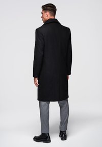 Manteau en laine noire avec un col noté, coupe ajustée et fente au dos. Associé à un pantalon gris et des bottines noires.