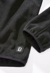 Svart fleeceplagg med strukturert overflate. Har elastiske mansjetter og en liten logoetikett på den nedre ermen.