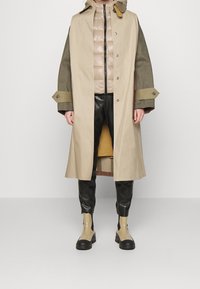 Beżowy płaszcz trench z zielonymi rękawami, pod spód położona beżowa kamizelka wypełniona, a na dole czarne syntetyczne spodnie. Całość dopełniają beżowo-czarne botki za kostkę.