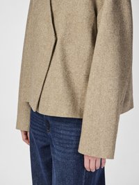 Jachetă bej din tweed cu un design structurat, având mâneci largi și o lungime ușor scurtă, asociată cu blugi din denim texturat de culoare albastru închis.