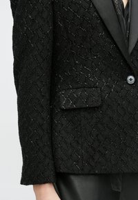 Zwart getextureerd colbert met een diamantenpatroon, voorzien van een satijnen revers, sluiting met één knoop en een klein borstzakje.