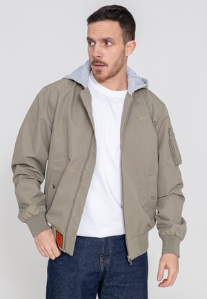 Licht khaki bomberjack met een grijze capuchon, zijzakken en ribgebreide manchetten. Voorzien van een ritssluiting en een opvallend oranje label.