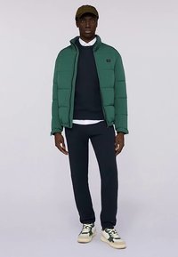 Zelená péřová bunda s vysokým límcem, tmavě modrý svetr, tmavě modré kalhoty a bílé tenisky se zelenými detaily, s texturovaným vzorem látky.