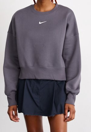Osoba nosząca szarą bluzę Nike z małym białym logo, w połączeniu z krótką czarną spódnicą, stojąca na jednolitym tle.