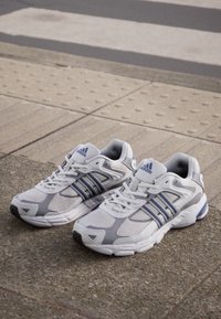 Des chaussures de sport blanches et grises en tissu respirant, avec des accents bleus et trois bandes latérales. Elles présentent une semelle rembourrée et un col rembourré.