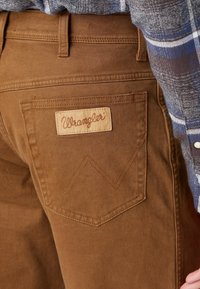 Bruna Wrangler-jeans bakficka med broderat "W"-mönster, bärs med en blå och grå rutad skjorta.