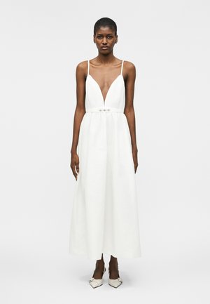 Γυναίκα που φοράει λευκό αμάνικο φόρεμα με βαθύ V ντεκολτέ και εφαρμοστή μέση, στέκεται μπροστά σε ένα απλό ανοιχτό φόντο.