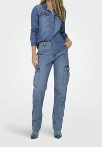 Frau in einem blauen Denim-Hemd, das in eine blaue Cargo-Jeans mit Taschen gesteckt ist, steht vor einem schlichten weißen Hintergrund.