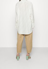 Chemise blanche à manches longues avec un motif texturé subtil, associée à un pantalon beige à jambe large et des sandales à talons blocs vert clair.