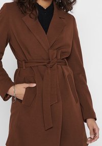 Mujer que lleva un abrigo marrón con cinturón sobre una blusa negra, con una mano en el bolsillo y pulsera y anillos de plata en las manos.