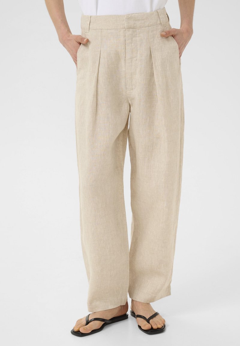 Personne portant un pantalon en lin beige à jambes larges, les mains dans les poches, et des sandales noires à brides sur fond blanc.