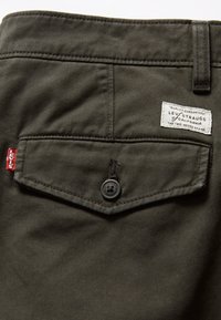 Gros plan sur la poche d’un pantalon Levi’s vert olive avec fermeture à bouton, étiquette rouge Levi’s, et étiquette blanche de la marque cousue au-dessus de la poche.