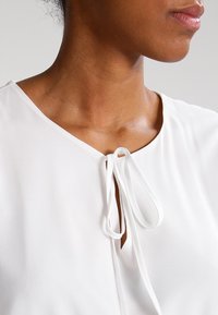 Valkoinen pusero, jossa on V-pääntie ja solmittava nauhakoriste, valmistettu sileästä kankaasta, joka hehkuu hienovaraisesti ja on pehmeästi teksturoitu.