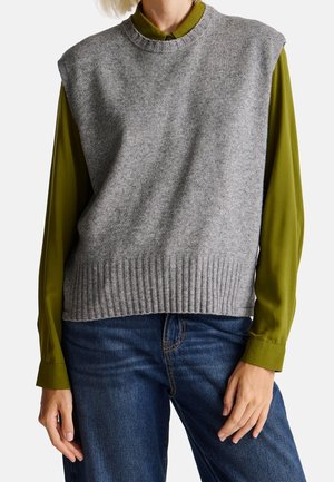 Personne portant un pull sans manches gris en maille par-dessus une chemise kaki à boutons et un jean bleu, debout les mains détendues.