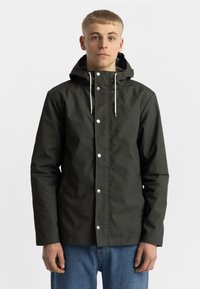Donkergroene, waterdichte jas met een capuchon, voorzien van witte drukknoopjes en beige trekkoorden. Draagt over blauwe jeans.