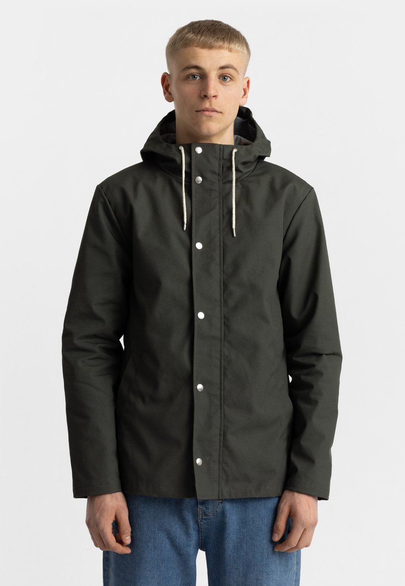 Donkergroene, waterdichte jas met een capuchon, voorzien van witte drukknoopjes en beige trekkoorden. Draagt over blauwe jeans.
