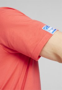 Koraalkleurig T-shirt met een zachte katoentextuur, voorzien van een logo-opdruk op de mouw en een relaxte pasvorm. Zichtbare stiknaaddetails.