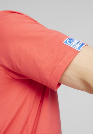 Korallefarbenes T-Shirt aus weichem Baumwollmaterial, mit einem Logopatch am Ärmel und einem entspannten Schnitt. Sichtbare Nahtdetails.