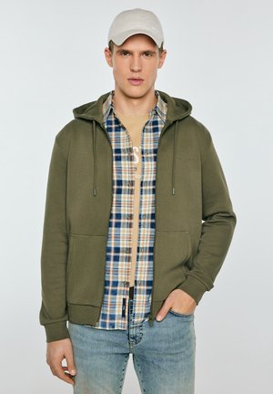 Młody mężczyzna w beżowej czapce, oliwkowozielonej bluzie z zamkiem, niebiesko-beżowej koszuli w kratę, beżowej koszulce i jasnoniebieskich dżinsach na jednolitym tle.