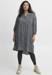 Laza fekete ruha zöld, kék és fehér virágmintával; V-nyakú és háromnegyedes ujjú. Fekete leggings-szel és bézs csizmával viselve.