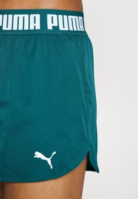 Shorts de sport teal avec un logo "PUMA" blanc sur la ceinture et un emblème de chat en saut. Présentent une texture lisse et des fentes latérales pour plus de flexibilité.