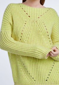 Pull tricoté vert citron avec une coupe ample, des poignets larges côtelés et des motifs de points ouverts décoratifs sur le devant. Le fil texturé apporte de la profondeur.