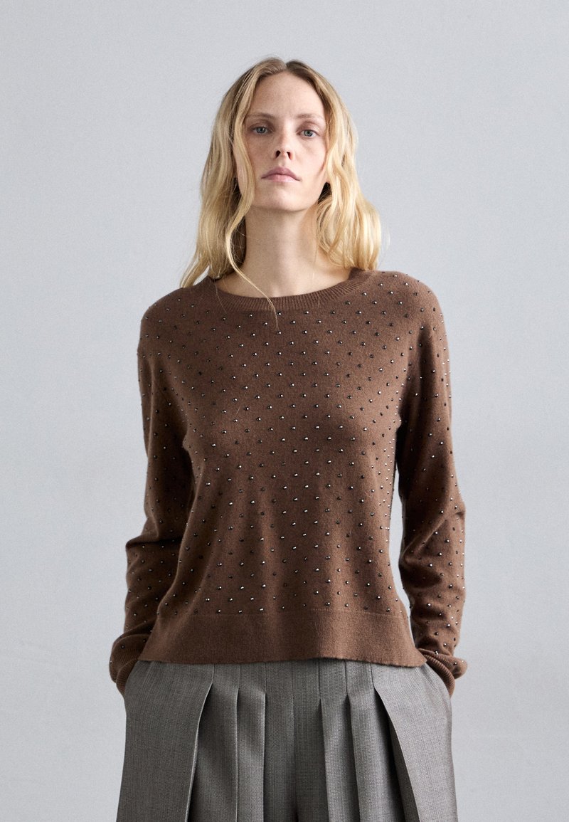 Kobieta z długimi blond włosami, ubrana w brązowy sweter z ćwiekami i szare plisowane spodnie, stojąca na tle gładkiego, jasnoszarego tła.