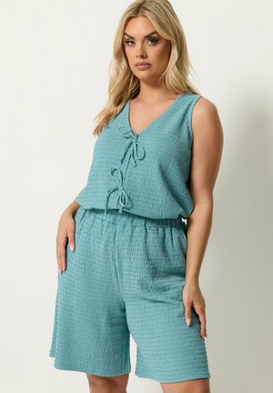 Blond kvinde står ved væggen iført ærmeløs tekstureret teal top med to frontbind og matchende elastisk talje knælange shorts.