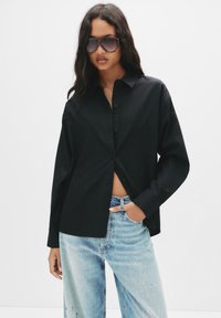 Camicia nera a maniche lunghe con bottoni, dal fit rilassato, abbinata a jeans chiari strappati. Il modello indossa occhiali da sole oversize.