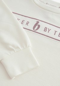 Šviesiai smėlinis ilgos rankovės džemperis su šonkačių tekstūra, turintis centrinius bordo spalvos grafikos juostą su užrašu "6 BY TED BAKER" ir sutrauktus rankogalius.