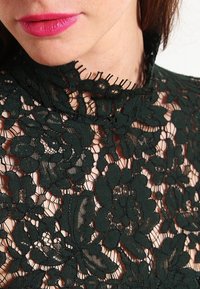 Haut en dentelle vert foncé avec des motifs floraux, col haut et bords effilochés ; la texture transparente révèle la peau sous le design complexe.