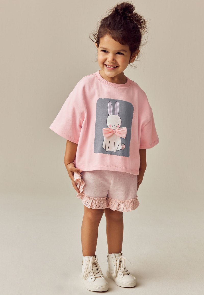 T-shirt oversize rose clair avec un motif de lapin et un nœud, assorti à un short rose clair avec bordure en dentelle, et des baskets montantes blanches.