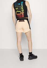Persona vistiendo una camiseta sin mangas negra con texto multicolor y pantalones cortos melocotón con paneles negros, calcetines negros y zapatos de senderismo negros, de pie de espaldas.