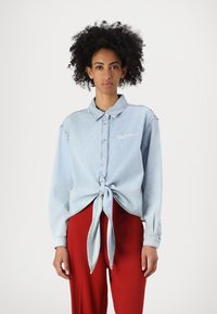 Ljusblå denimskjorta med knappar, långa ärmar och knutdetalj fram, med vit logotypbrodyr och en klassisk krage.