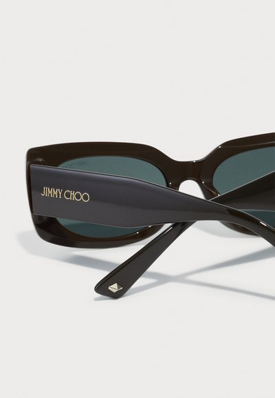 Ochelari de soare negri cu ramă unghiulară și lentile verzi. Text „JIMMY CHOO” aurit pe lateral. Brațe negre texturate cu un detaliu metalic.