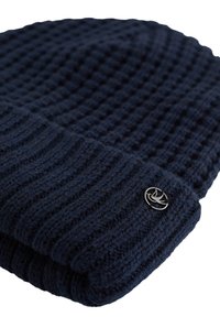 Mornarsko pletena kapa z teksturiranim rebrastim vzorcem, ki vključuje srebrni logotip, vezen na zloženem robu.
