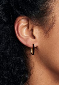 Czarno-złote kolczyki w kształcie obrączek. Obrączki są cienkie, mają gładką teksturę oraz kontrastowy owalny kształt. Noszone na uchu.