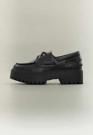 Sort læder platform loafer med chunky rillede sål og snøredetaljer på glat neutralt baggrund.