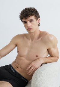 Zwarte Calvin Klein boxer shorts op een shirtloze jonge man met krullend haar, zittend op een licht gestructureerde stoel tegen een effen achtergrond.