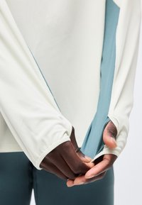 Käed tõmbavad valge ja sinise tekstuuriga pika varrukaga sportliku särgi mansetti, millel on pöidlate jaoks augud, kantud tumedate pükste peal.