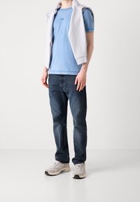 Jasnoniebieska bawełniana koszulka z małym logo, w połączeniu z ciemnoniebieskimi dżinsami i szarymi sneakersami. Biała bluza jest zarzucona na ramionach.