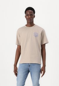 Beige bomull t-skjorte med rund hals og korte ermer, med et blått trykt design på venstre bryst. Klassisk passform, myk tekstur.
