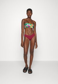 Biquíni com um soutien de estampado tropical multicolorido e partes de baixo em vermelho escuro de corte baixo. Modelo usa sandálias pretas de slide. Tecido suave, design ajustado.