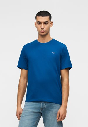 Jeune homme portant un t-shirt bleu uni à manches courtes avec un petit texte blanc sur la poitrine et un jean bleu clair, posant devant un fond blanc.