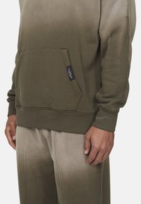Felpa verde oliva con tasca frontale, polsini a coste e pantaloni della tuta grigi chiari sbiaditi, caratterizzati da una texture a gradiente e cuciture visibili.
