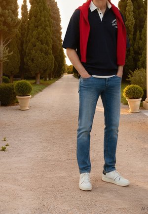 Personne debout sur un chemin de gravier entre de grands arbres, portant un jean bleu, des baskets blanches, un polo noir et un pull rouge posé sur les épaules.