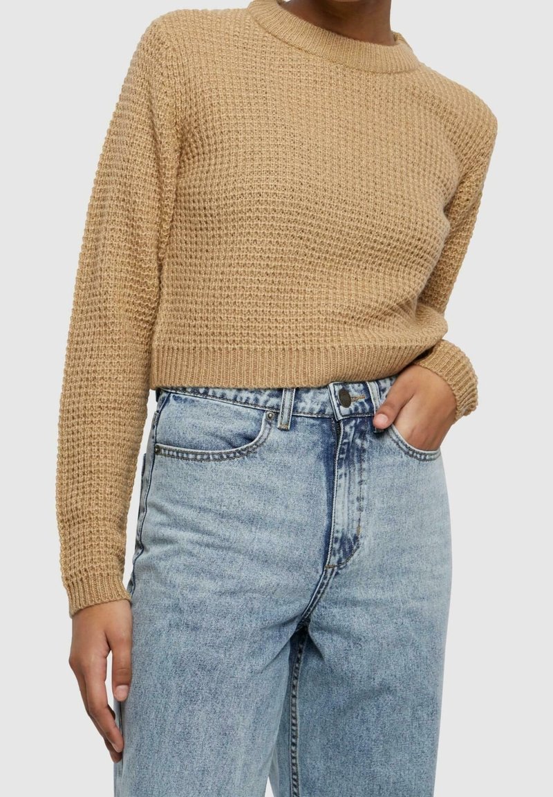 Personne portant un pull en maille beige texturé rentré dans un jean taille haute bleu clair, une main dans la poche, sur un fond neutre.