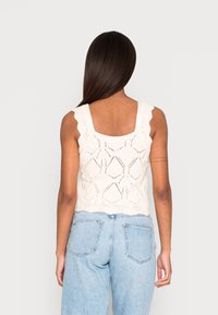 Pletený krémový top na ramínka s vlnitými popruhy, zdobený geometrickými vzory a texturovanými detaily, doplněný o světle modré džíny s vysokým pasem.