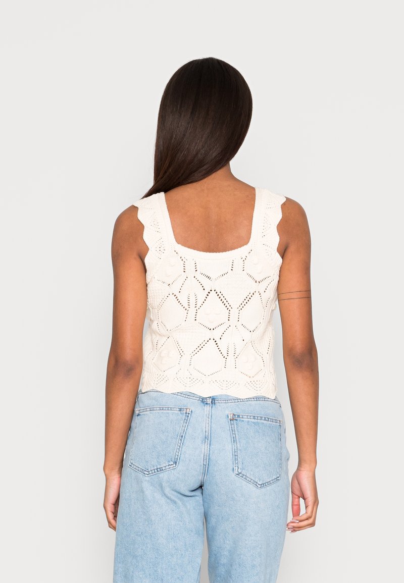 Pletený krémový top na ramínka s vlnitými popruhy, zdobený geometrickými vzory a texturovanými detaily, doplněný o světle modré džíny s vysokým pasem.