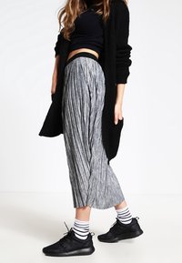 Žena oblečená v černém svetru, černém krátkém topu, šedé skládací sukni, pruhovaných ponožkách a černých teniskách, postávající na bílém pozadí.